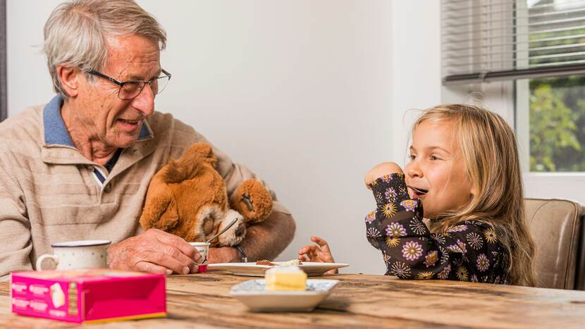 opa zit met kleindochter aan een tafel, opa houdt een knuffelbeer vast, kleindochter eet een dessertje