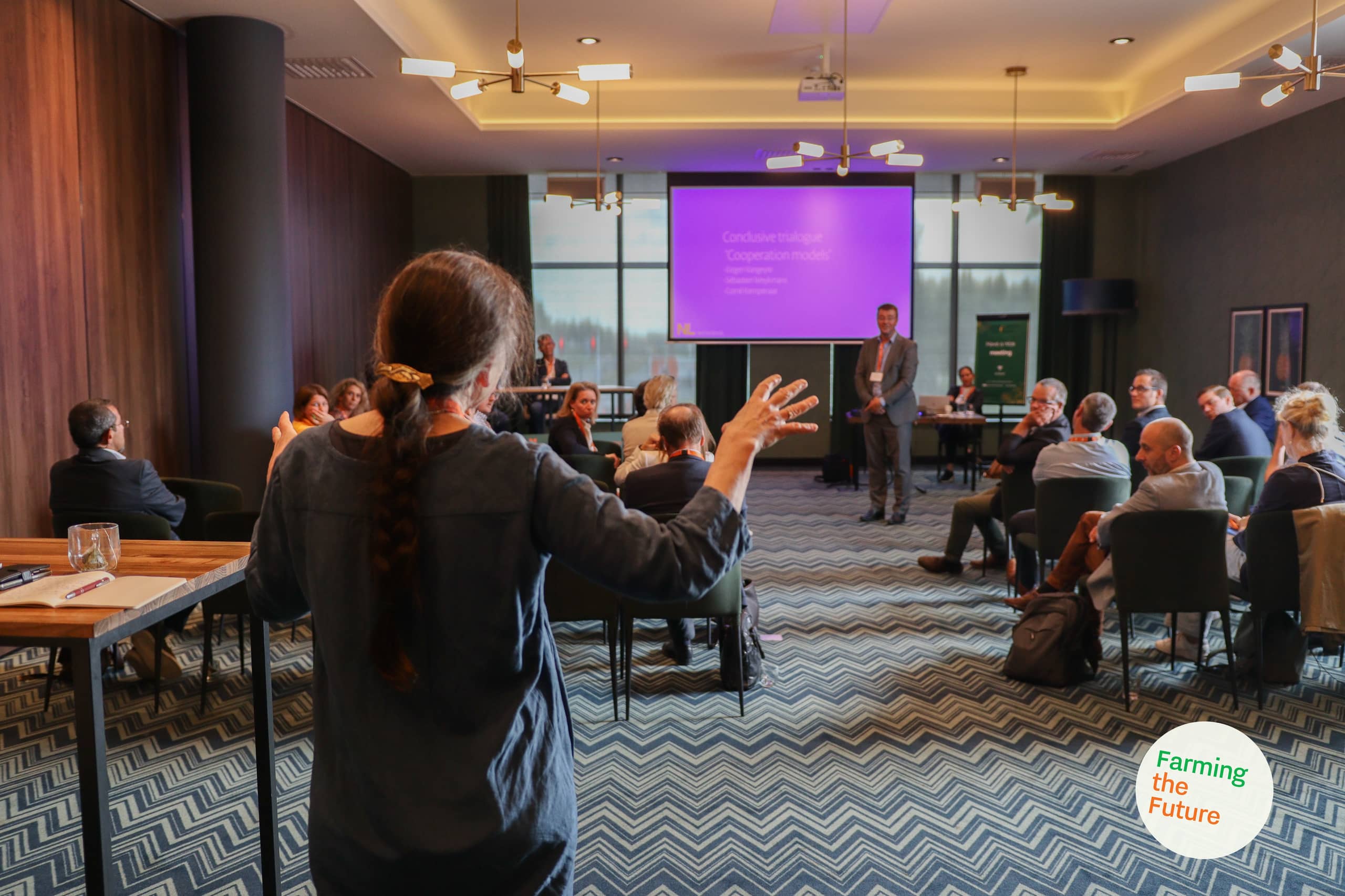 Links op de voorgrond staat een vrouw die spreekt tijdens discussie in een vergaderzaal van een hotel, op de achtergrond zitten mensen te luisteren, terwijl de moderator voor een groot projectiescherm staat