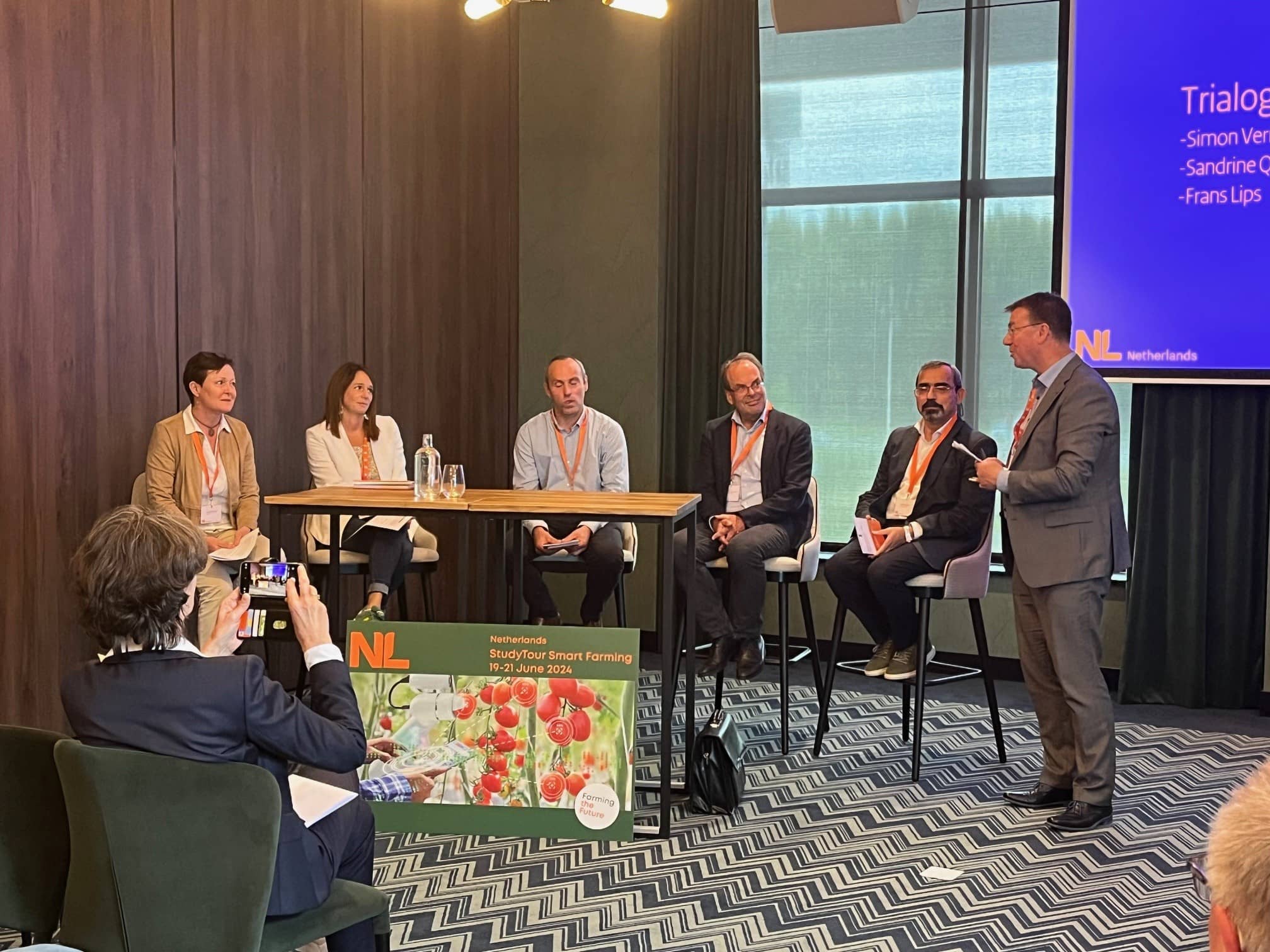 Vijf mensen zitten in een vergaderzaal van een hotel op een hoge kruk met de moderator naast hen. Voor hen staat een tafel met wat glaasjes water.