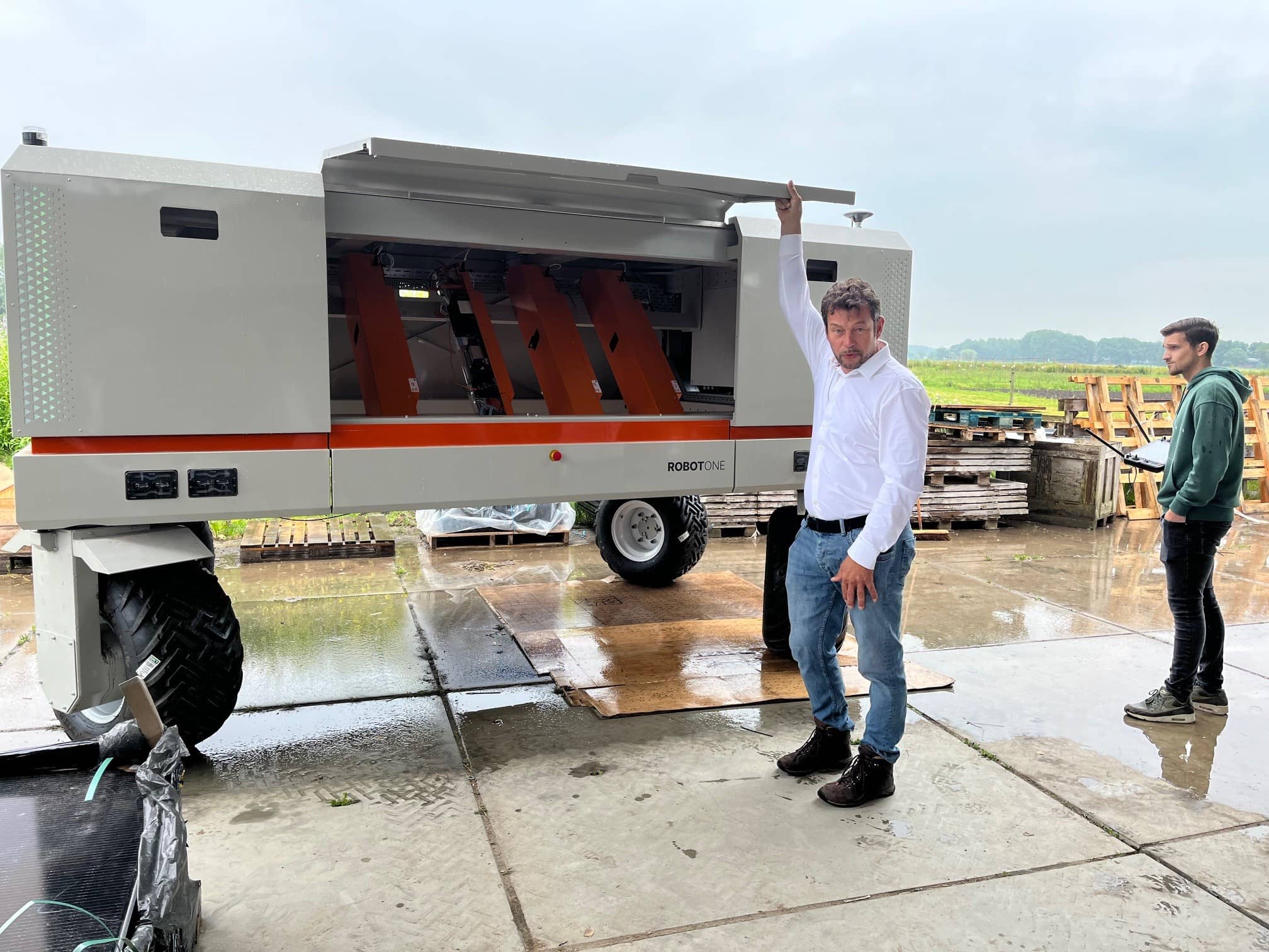 Man die voor een agrarische robot staat en met zijn rechterarm een klep van de robot open houdt