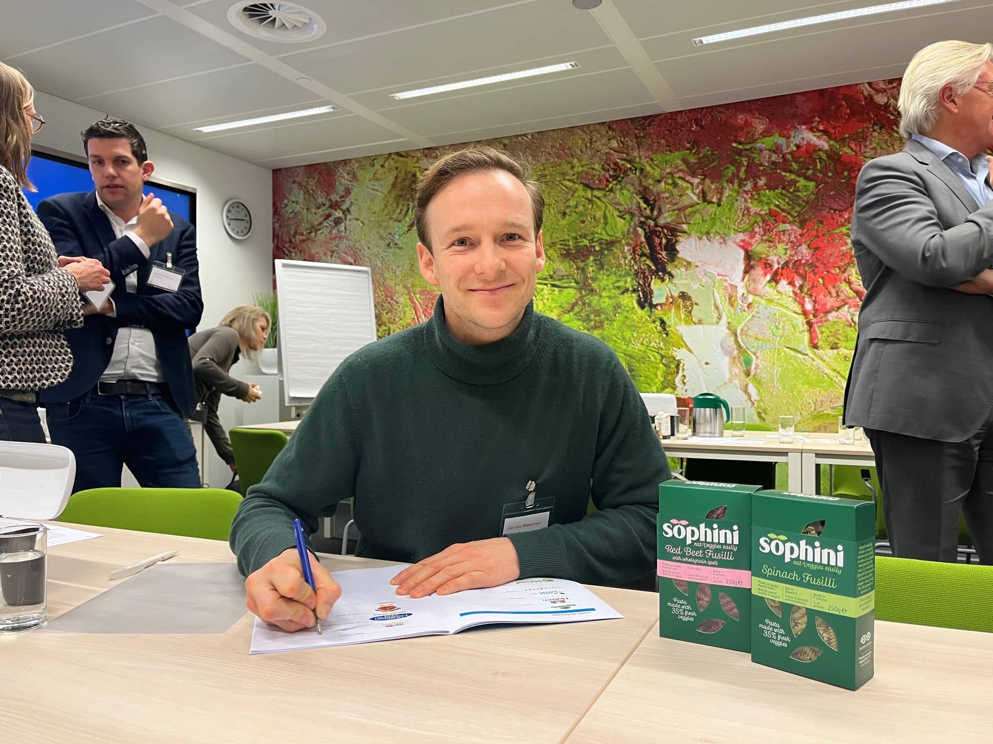 Jonge man van het pastamerk Sophine zit achter een tafel en kijkt in de lens terwijl hij een pen vasthoudt om een document te ondertekenen