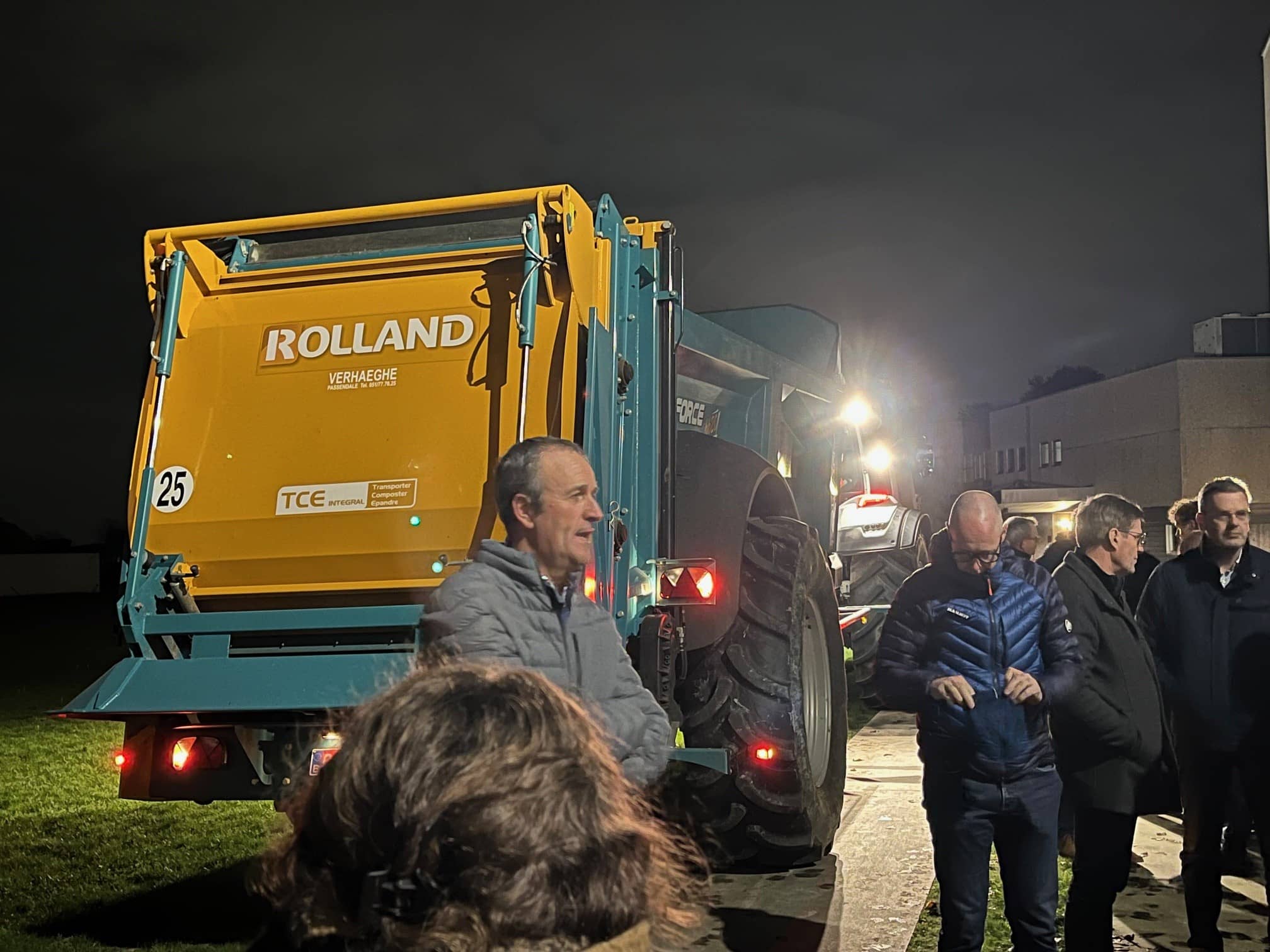 een landbouwer staat bij zijn tractor met aanhanger en vertelt terwijl een groep mensen luistert