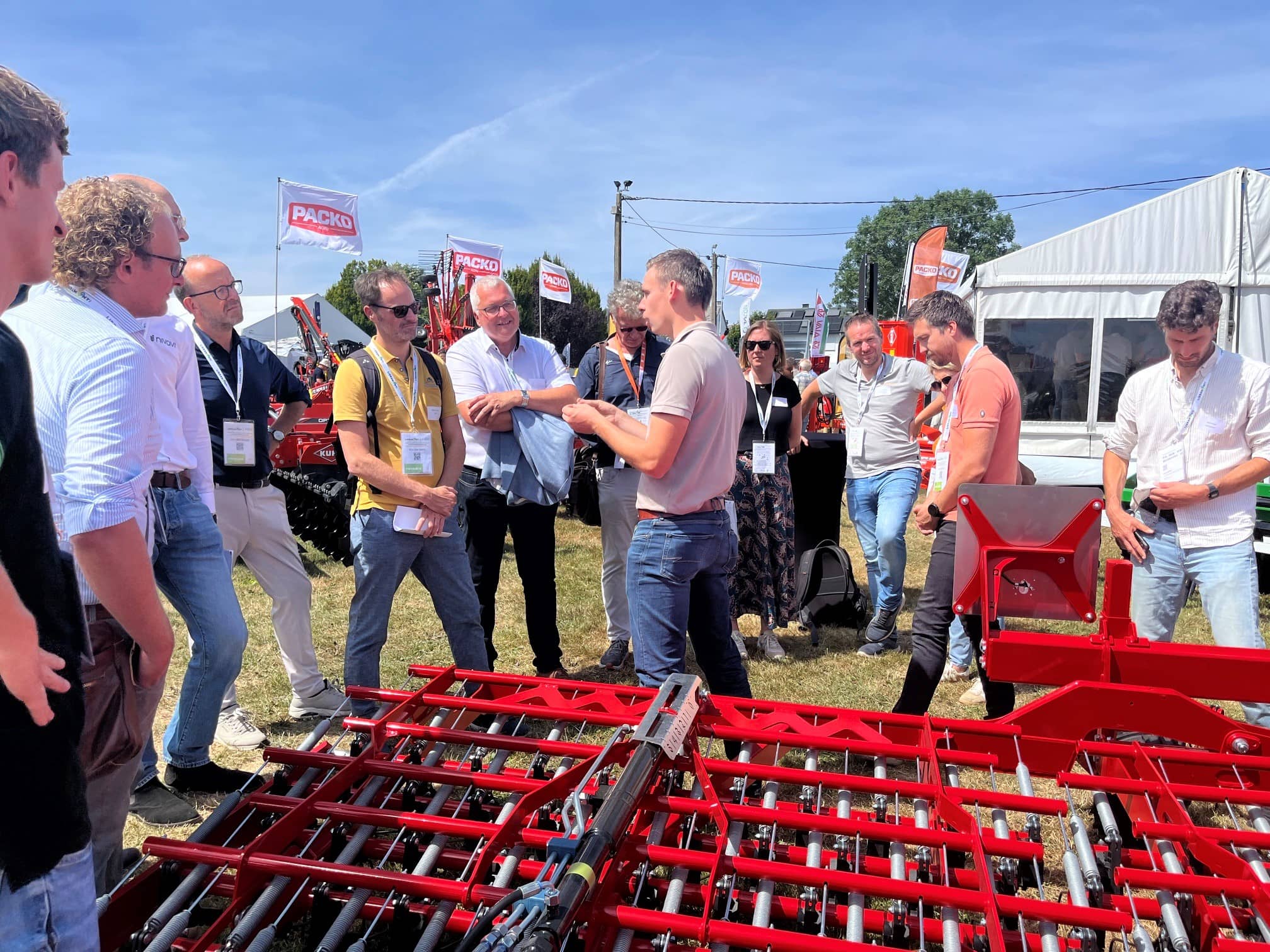 een groep mannen staat op een beurs bij een grote landbouwmachine en luistert naar een uitleg over de machine