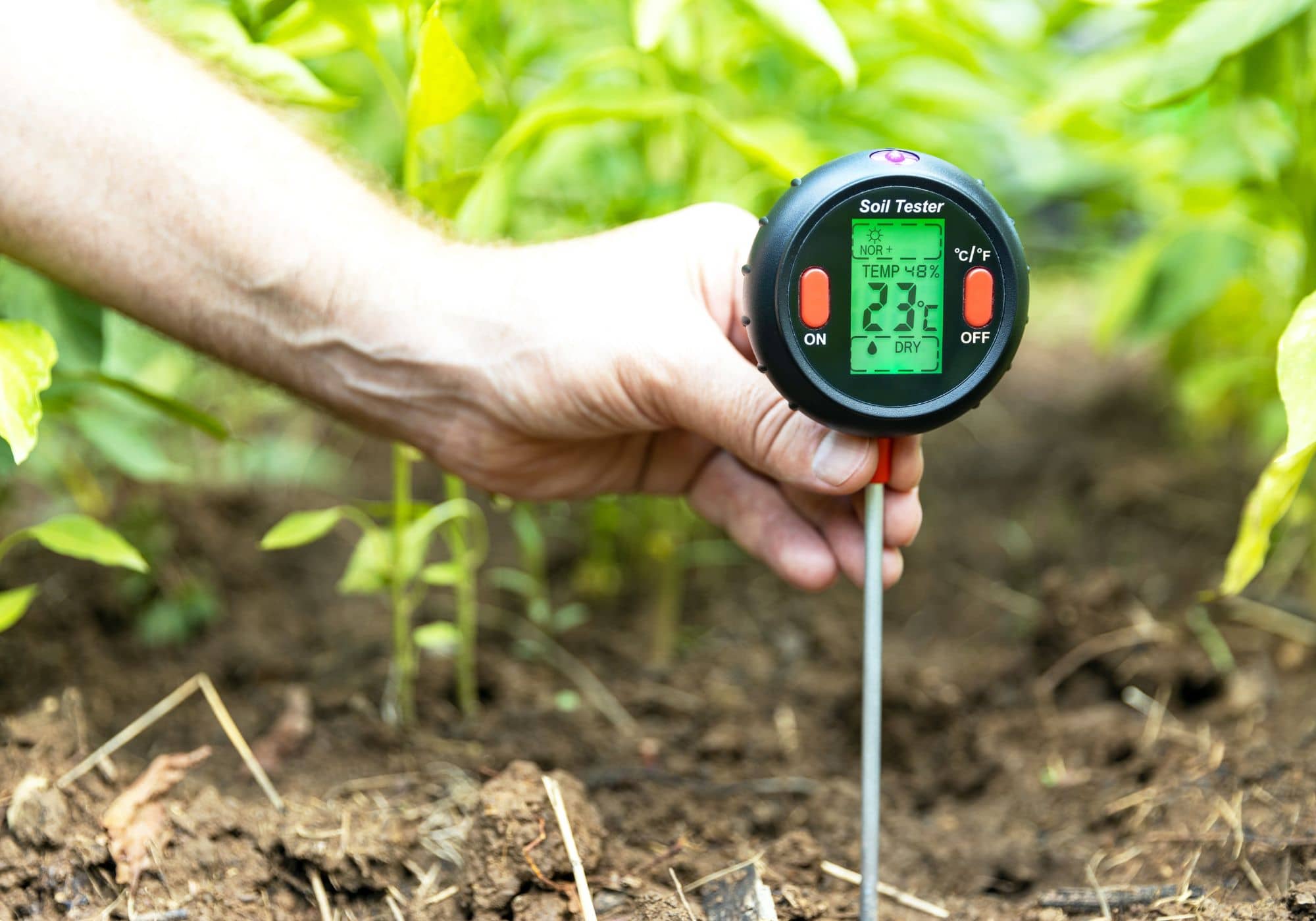een hand houdt een sensor vast die in de grond van een akker steekt, de sensor geeft o.a. de bodemvochtigheid weer