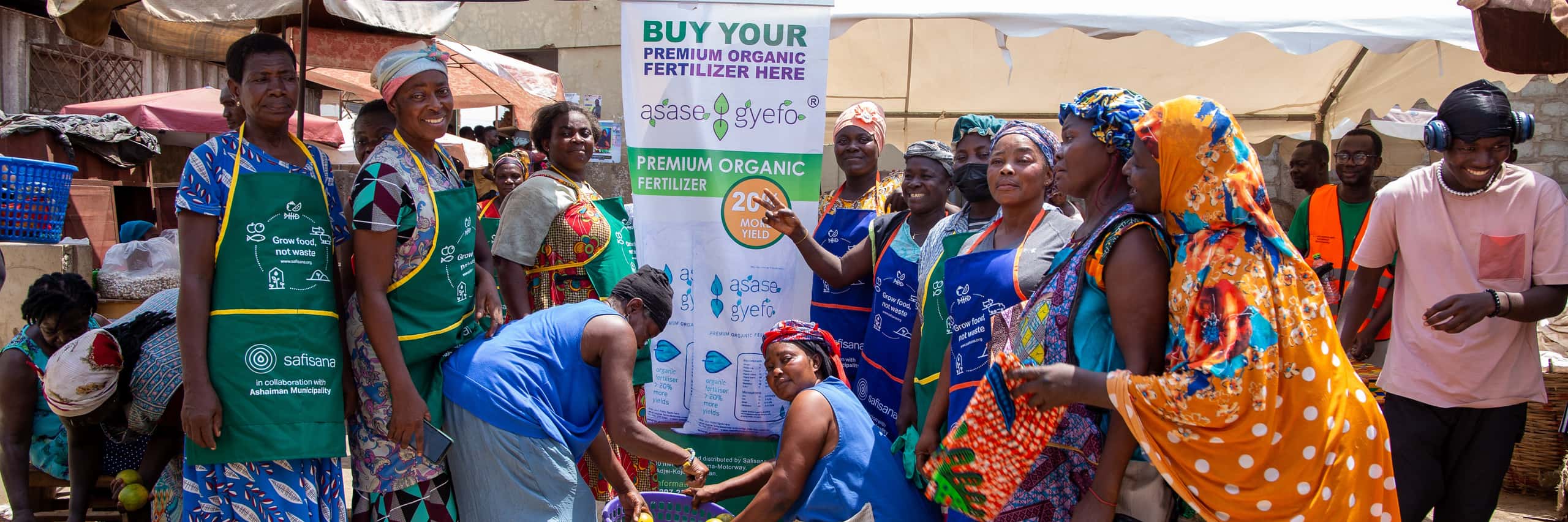 a group of women stand beside a Asaase Gyefo banner at a market