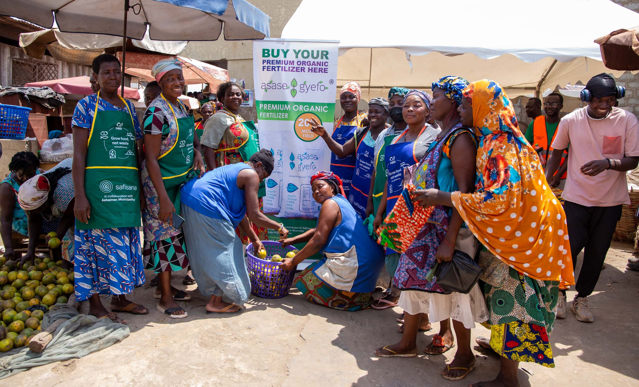a group of women stand beside a Asaase Gyefo banner at a market