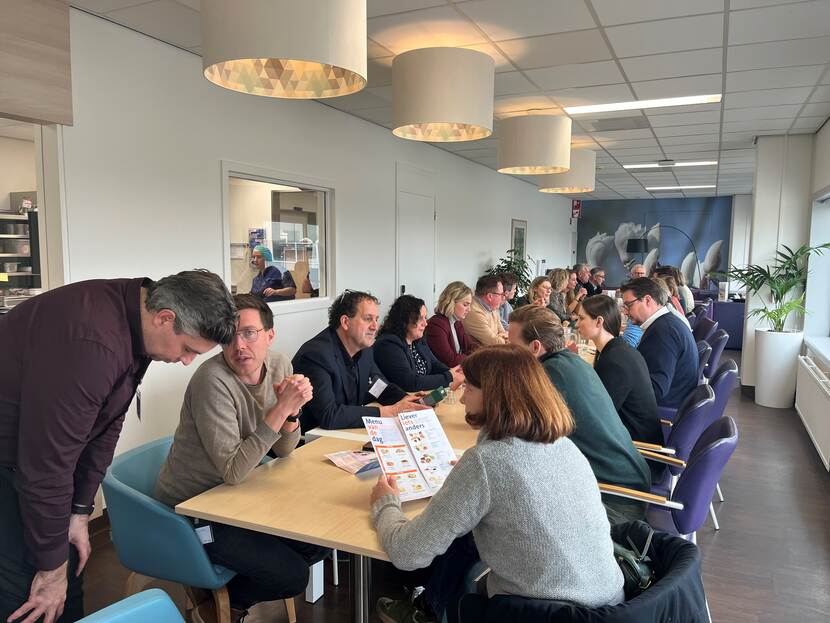 Groep van mensen zittend aan een lange tafel in de bistro van het Universitair Medisch centrum Maastricht, met lunchkaart voor hen
