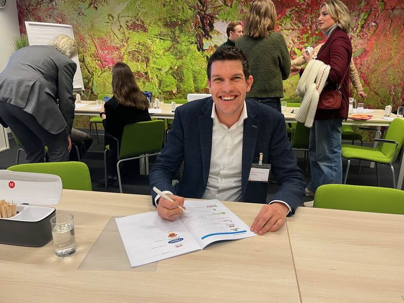 Man kijkt lachend in de lens terwijl hij achter een tafel zit met een pen in zijn hand, klaar om een document te ondertekenen.