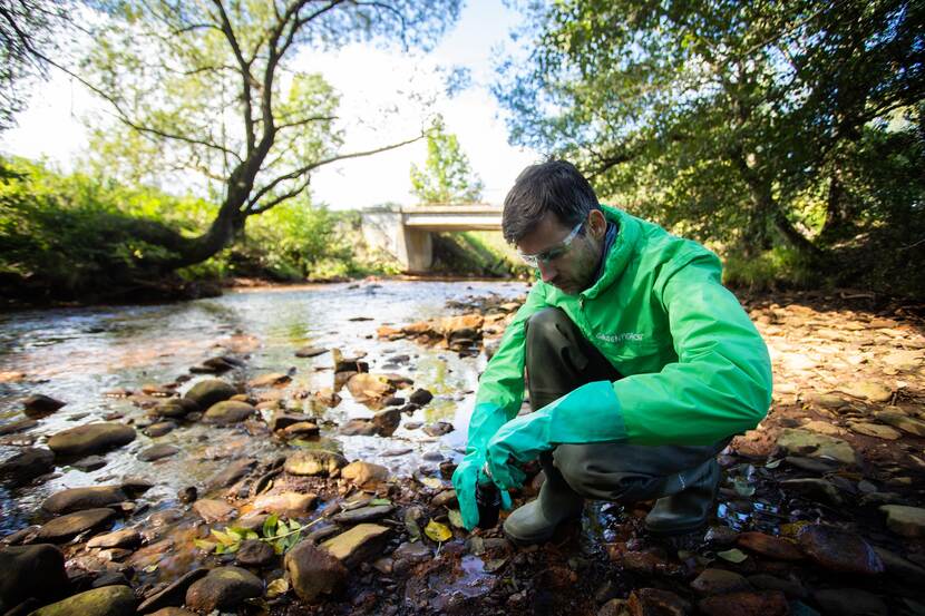 A person from Greenpeace, wearing a plastic protective suit of some sort and safety goggles, is taking a sample from the water at a shallow section of the River Sajó.
