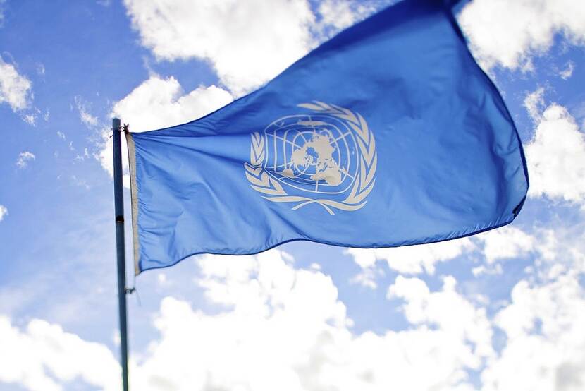 The flag of the United Nations rippling and waving in the wind against the backdrop of a bright blue sky dotted with white clouds.