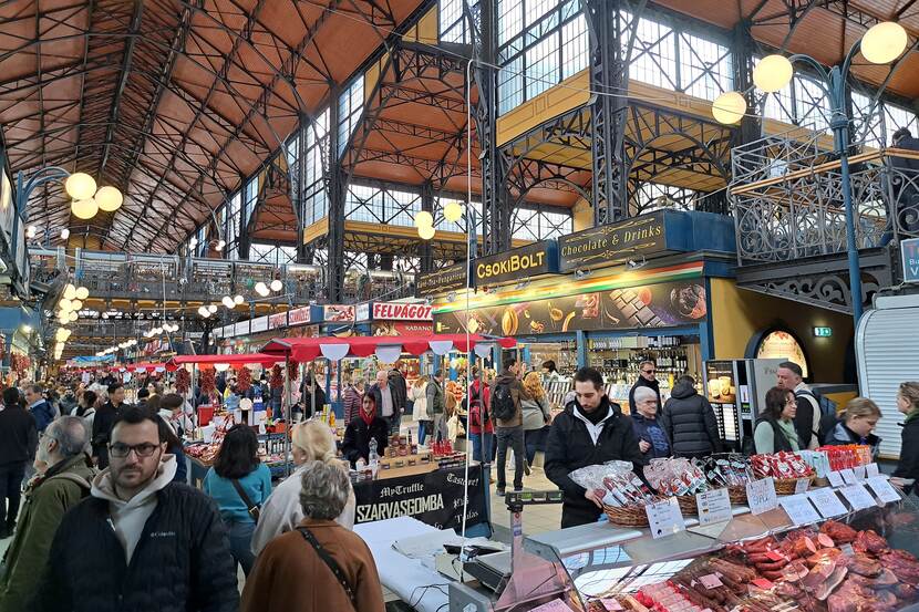 Busy market hall in Budapest. The hall is built in Victorian industrial style, with impressive, arching steel beams supporting a high ceiling and steel-reinforced high windows letting in natural light. People are passing by stalls and large booths, with various products on display.