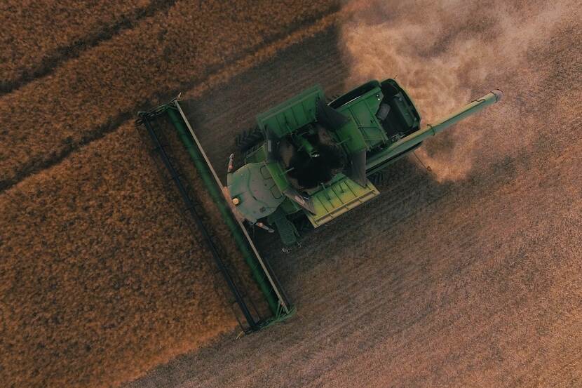 In a close-up aerial shot, a combine harvester can be seen, harvesting a field of wheat or some other cereal. Ambient lighting makes the photo more dramatic, suggesting that it is late afternoon. There is a trail of dust behind the machine as it is mowing down stalks of crops, leaving behind it an empty field.