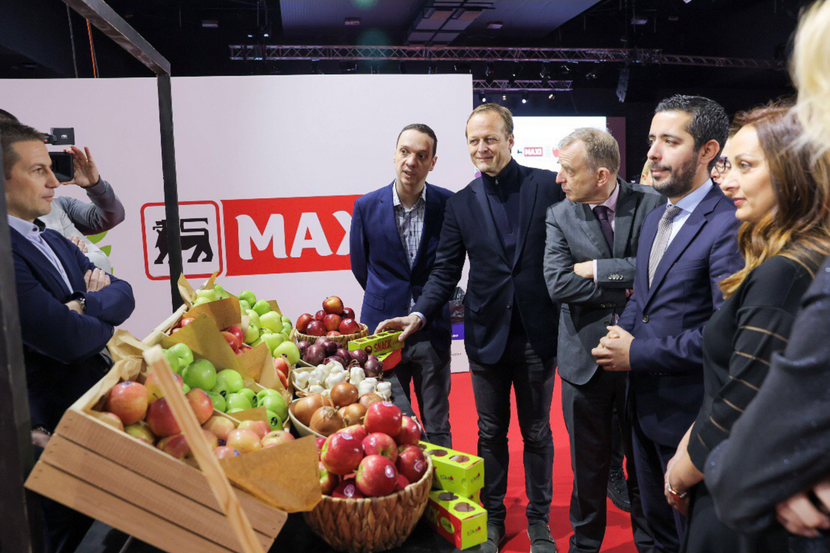 A stand at a food expo. The farmer who owns the stand stands behind it with arms folded. High-profile guests posein front of the stand which is full of fresh fruits. Everyone is wearing suits.