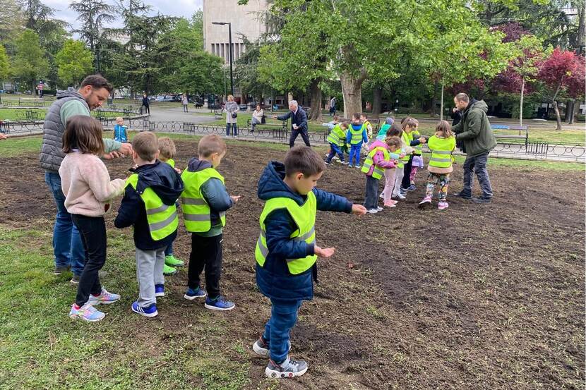 A group of children wearing visibility vests are planting seeds in a park under the supervision of two adults.