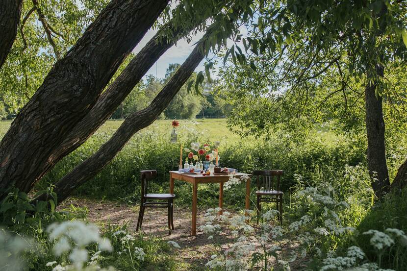 A table with flowers in vases can be seen under trees, with two chairs on each side. It is summer, the photo has an air of bucolic tranquility.