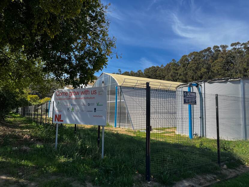 Image of a greenhouse situated behind a fence. To the left is a sign with logos of companies who have built the greenhouse, and to the right is a rainwater tank.